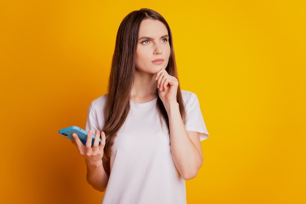 Photo de dame d'esprit tenir le téléphone pense regarder l'espace vide porter un t-shirt blanc posant sur fond jaune