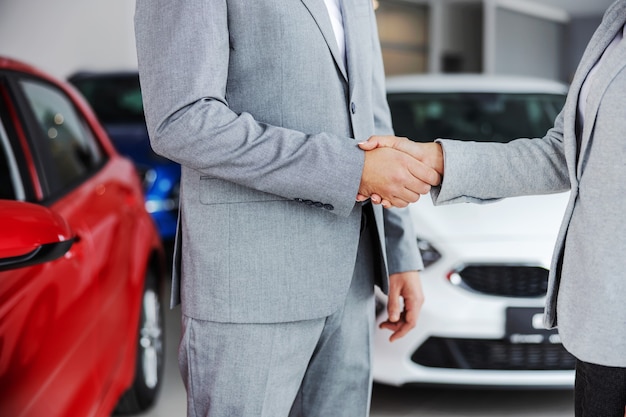 Photo découpée du vendeur de voiture serrant la main de l'acheteur en se tenant debout dans le salon de l'automobile.