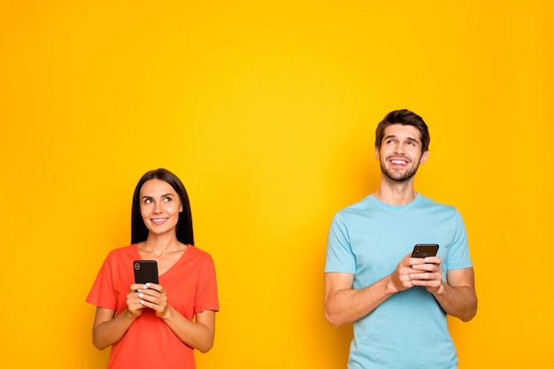 Photo de deux drôles de gars dame personnes couple tenir téléphone bras regardant un espace vide ont idée de texte post créatif porter des t-shirts orange bleu décontracté mur de couleur jaune isolé