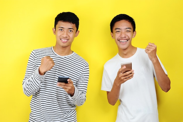 Photo de deux hommes asiatiques excités souriants à l'aide de téléphones portables isolés sur fond jaune
