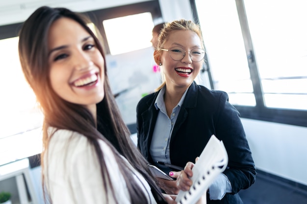 Photo de deux jeunes femmes d'affaires élégantes souriant et regardant la caméra dans un lieu de coworking.