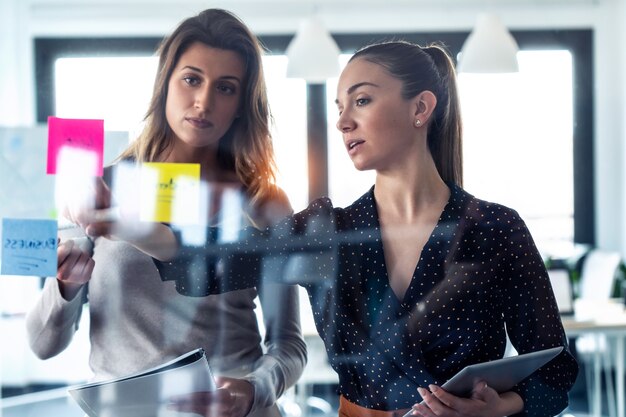 Photo de deux jeunes femmes d'affaires travaillant ensemble sur du verre mural avec des autocollants post-it sur un espace de coworking.