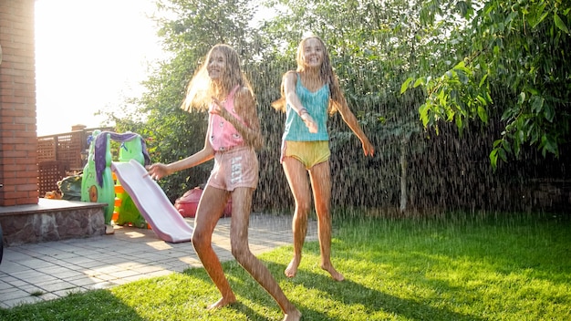 Photo de deux soeurs riantes heureuses dans des vêtements mouillés dansant sous les gouttelettes d'eau du tuyau d'arrosage au jardin. Famille jouant et s'amusant à l'extérieur en été