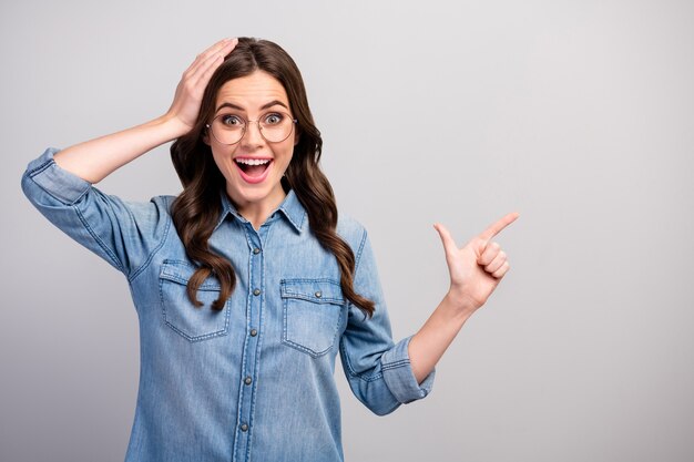 Photo de drôle de femme d'affaires bouche ouverte indiquer l'espace vide de doigt conseiller produit nouveauté bras sur la tête porter des spécifications jeans casual chemise en jean couleur gris isolé