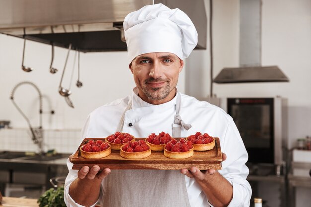 Photo du chef de sexe masculin adulte en uniforme blanc tenant la plaque avec des gâteaux