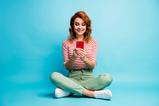 Photo du corps entier d'une femme joyeuse positive s'asseoir les jambes croisées utiliser smartphone lire partager les nouvelles des médias sociaux porter des vêtements élégants isolés sur la couleur bleue