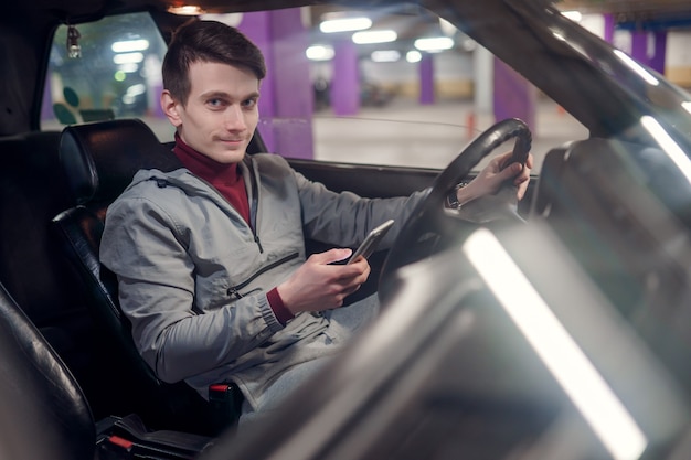 Photo du côté d'un conducteur masculin souriant avec un téléphone dans les mains assis dans une voiture sur une assurance souterraine