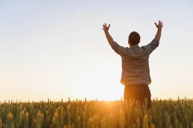 Photo du lever ou du coucher du soleil d'un gars avec les mains levées regardant le soleil et profitant de la journée Homme adulte debout seul au milieu d'un champ de blé mûr