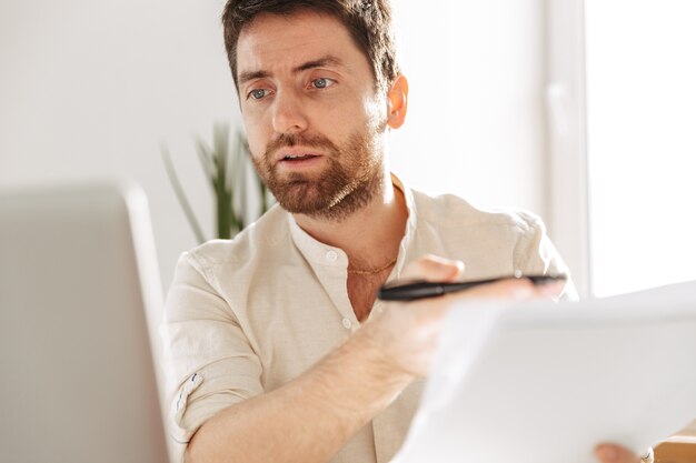 Photo d'un employé de bureau concentré de 30 ans portant une chemise blanche à l'aide d'un ordinateur portable et de documents papier, assis à table dans un lieu de travail moderne