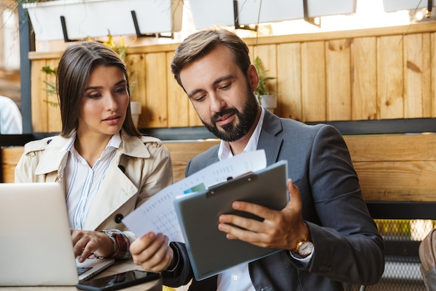 Photo d'employés de bureau concentrés homme et femme en tenue de soirée regardant des documents tout en utilisant un ordinateur portable et un téléphone portable au café