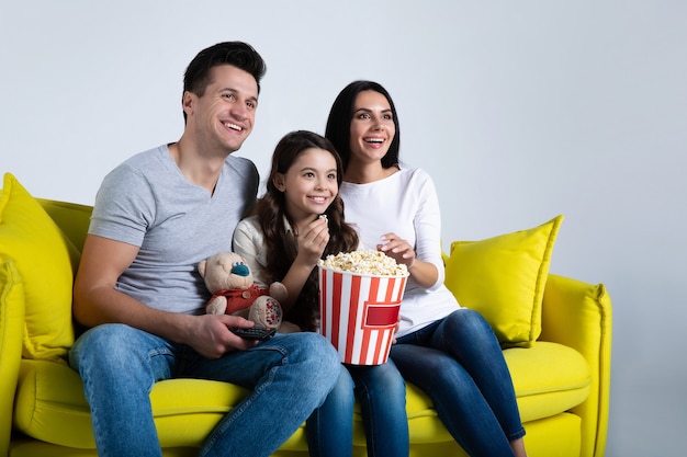 Photo d'un enfant mignon et de ses parents, qui mangent du pop-corn ensemble, tout en regardant des émissions de télévision sur le canapé.