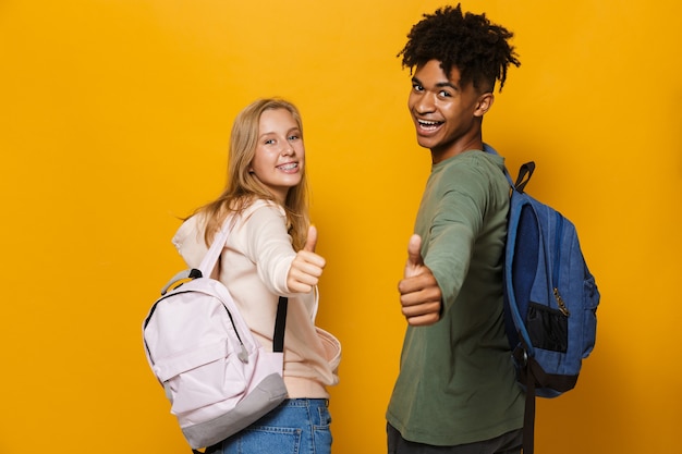 Photo d'étudiants heureux homme et femme de 16 à 18 ans portant des sacs à dos riant et montrant les pouces vers le haut, isolés sur fond jaune