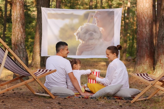 Photo d'une famille assise dans la forêt sur des chaises longues avec leur petite fille et regardant un film sur un projecteur parents se regardant avec le sourire famille profitant du temps passé ensemble