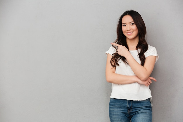 Photo de femme affable asiatique en t-shirt décontracté et jeans posant sur l'appareil photo et pointant le doigt de côté sur le fond, isolé sur le mur gris