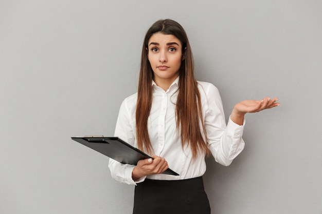 Photo de femme d'affaires en chemise blanche et jupe noire tenant le presse-papiers avec des documents au bureau et faisant des gestes de la main dans la perplexité, isolé sur mur gris