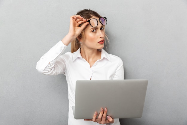 Photo de femme d'affaires européenne portant des lunettes debout et tenant un ordinateur portable au bureau, isolé