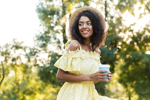 Photo d'une femme afro-américaine de 20 ans portant un chapeau de paille, marchant en plein air et tenant un café à emporter