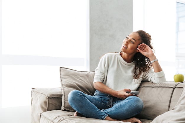 Photo D'une Femme Afro-américaine Heureuse à L'aide De Téléphone Portable, Assis Sur Un Canapé Dans Un Appartement Lumineux