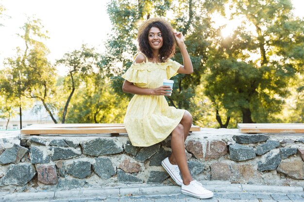 Photo d'une femme américaine de 20 ans portant un chapeau de paille et une robe, assise sur un banc dans un parc et buvant du café à emporter