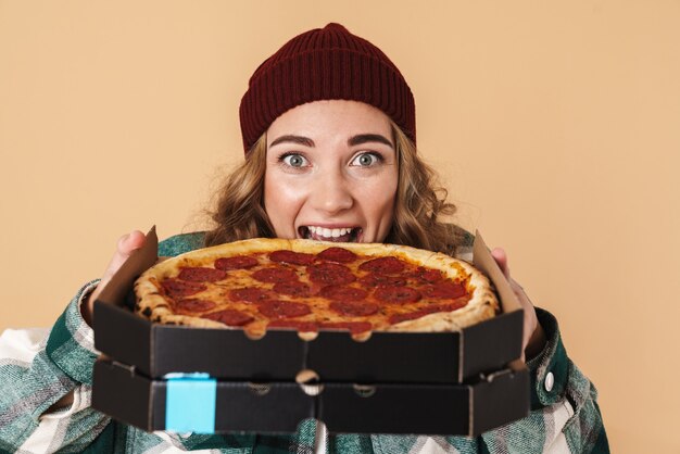 Photo photo d'une femme assez excitée en bonnet tricoté mangeant une pizza isolée sur beige