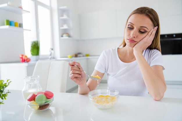 Photo de femme au foyer assez bouleversée tenant une cuillère ne veulent pas manger de cornflakes de petit déjeuner de lait fatigué de suivre un régime s'ennuie assis table cuisine lumière blanche à l'intérieur