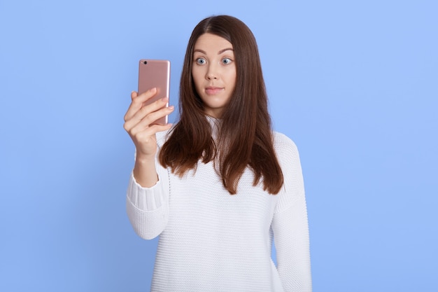 Photo d'une femme aux cheveux noirs surpris porte un pull blanc, tient un téléphone mobile, reçoit un message, regarde avec une expression choquée à l'écran du téléphone portable, pose isolée à l'intérieur