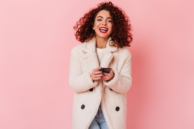 Photo d'une femme bouclée positive avec des lèvres brillantes riant sur fond rose Fille en blouse blanche tenant un smartphone