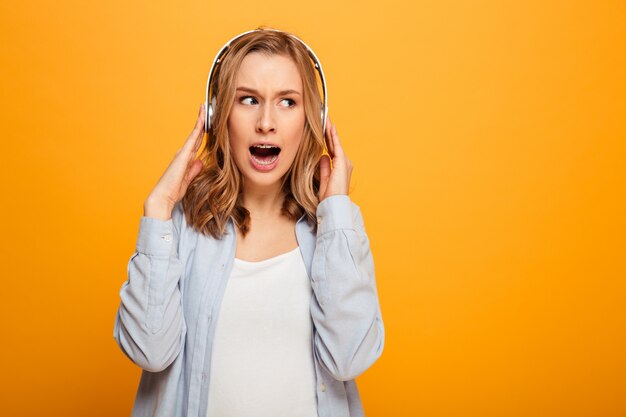 Photo d'une femme brune portant des bretelles réagir émotionnellement sur la musique tout en écoutant une chanson ou un morceau à l'aide d'un casque sans fil, isolé sur un espace jaune