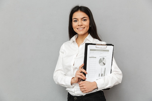 Photo de femme de bureau souriant portant une tenue professionnelle tenant le presse-papiers avec des papiers et à la caméra, isolé sur fond gris