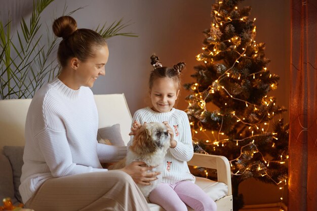 Photo d'une femme avec une coiffure chignon portant un chandail blanc assise avec sa petite fille et jouant avec leur chien pékinois s'amusant pendant le réveillon du Nouvel An