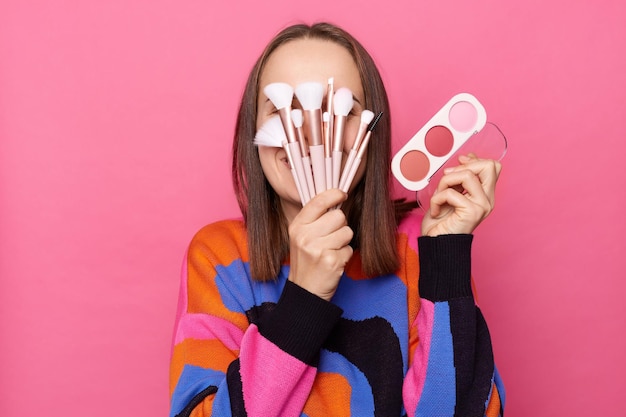 Photo d'une femme drôle anonyme avec des cheveux raides foncés portant un cavalier debout isolé sur fond rose couvrant son visage avec des pinceaux cosmétiques tenant une palette de fards à paupières