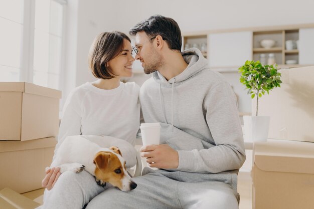 Photo d'une femme et d'un homme charmants qui vont embrasser leur mari tient un café à emporter assis à l'intérieur près de boîtes en carton déballent leurs effets personnels le jour du déménagement vivent avec un animal de compagnie acheter un nouvel appartement coûteux