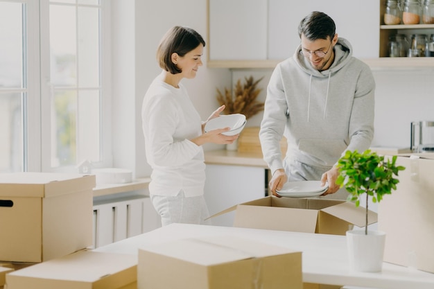 Photo d'une femme et d'un homme mariés heureux déballant la vaisselle des boîtes habillées avec désinvolture tenant des assiettes posées dans une nouvelle cuisine moderne entourée de colis mobiles Concept de maison et de réinstallation des personnes