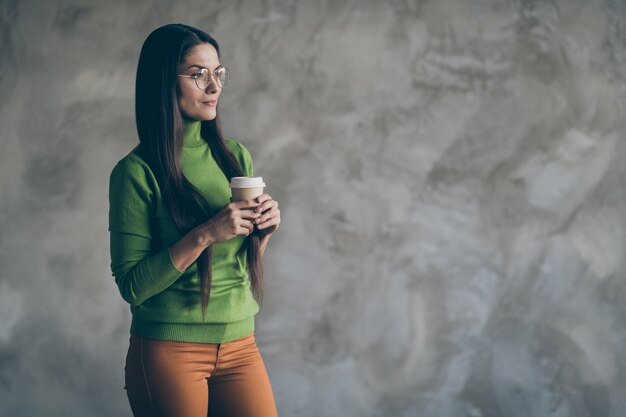 Photo de femme intéressée pensif pensif tenant tasse de boisson chaude en pantalon pantalon ornage à la recherche dans un espace vide mur de couleur gris isolé fond béton