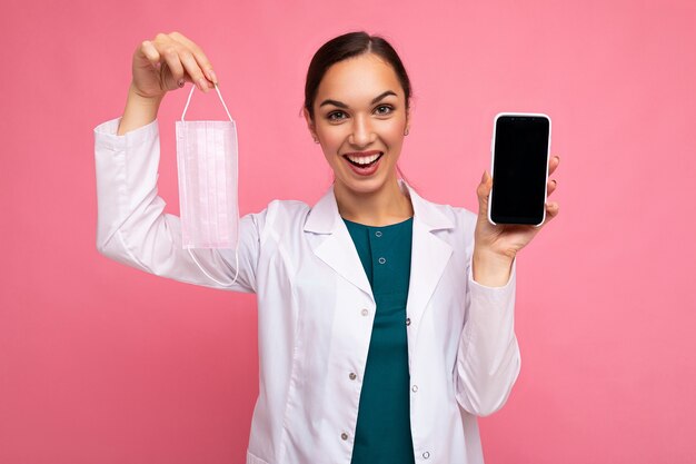 Photo d'une femme médicale adulte souriante portant un manteau blanc tenant un masque montrant un téléphone portable moderne isolé sur fond rose regardant la caméra. Maquette, découpe