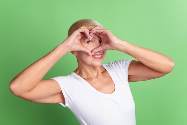 Photo d'une femme mûre gaie montre les doigts symbole du coeur des sentiments de cupidon rêve isolé sur fond de couleur verte.