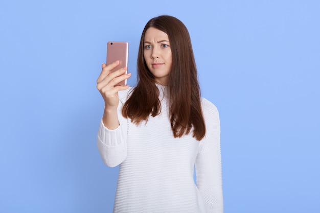 Photo d'une femme sérieuse utilise un appareil moderne, parcourt les médias sociaux, porte des vêtements décontractés, isolé sur un mur bleu