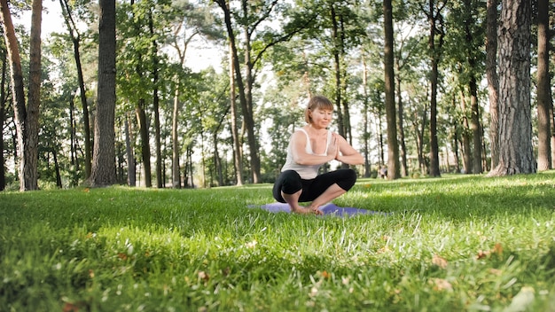 Photo d'une femme souriante d'âge moyen pratiquant le yoga et méditant au parc. Femme s'étirant et faisant du fitness sur tapis à forest