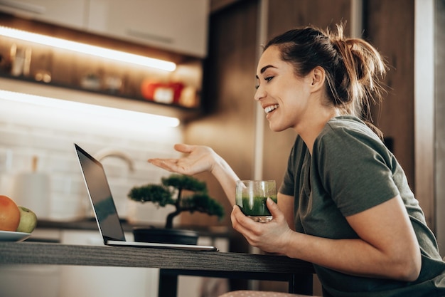 Photo D'une Femme Utilisant Son Ordinateur Portable Et Prenant Son Petit-déjeuner Dans Sa Cuisine Le Matin à La Maison.