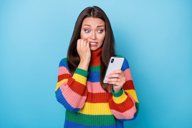 Photo photo d'une fille de blogueuse effrayée qui utilise un smartphone pour lire des nouvelles horribles sur les médias sociaux mordant les doigts les dents portent un pull de style arc-en-ciel isolé sur un fond de couleur bleu