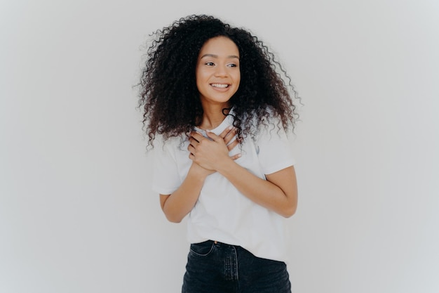 Photo d'une fille heureuse garde les deux mains sur la poitrine regarde de côté avec un sourire agréable remarque quelque chose de génial pose sur fond blanc porte un t-shirt décontracté et un jean Concept d'émotions positives