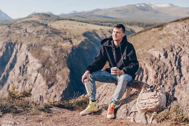 Photo avec un filtre. Un jeune homme vêtu d'un sweat à capuche noir regarde le paysage montagneux envoûtant tenant un thermos et une tasse de voyage blanche dans ses mains. Il y a un sac à côté.