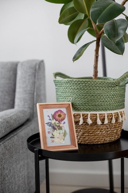 Une photo de fleurs séchées dans un cadre en bois à l'intérieur sur une table près d'un canapé gris et d'une maison