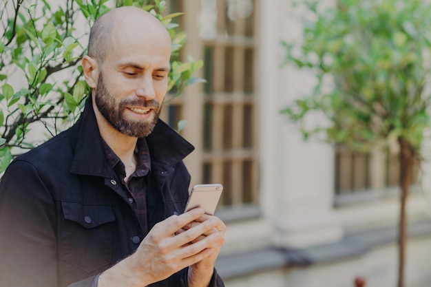 Photo franche d'un homme barbu chauve d'âge moyen a un sourire positif regarde attentivement le téléphone intelligent lit les informations nécessaires dans les poses Internet sur fond flou avec des arbres verts