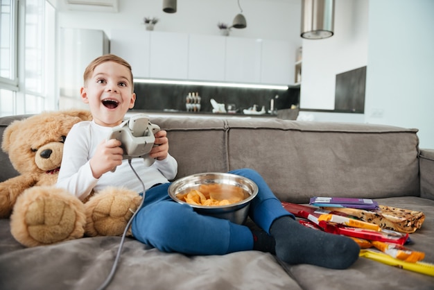 Photo d'un garçon joyeux sur un canapé avec un ours en peluche à la maison jouant à des jeux sur console tout en mangeant des chips. Manette de maintien.