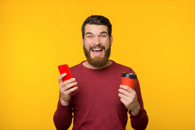 Photo d'un gars barbu excité en pull rouge, souriant et regardant la caméra tout en tenant son téléphone portable et une tasse de café