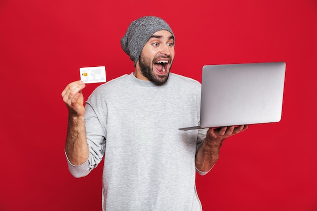 Photo de gars optimiste de 30 ans en tenue décontractée tenant une carte de crédit et un ordinateur portable en argent isolé