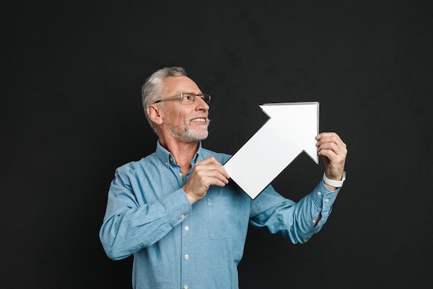 Photo de gentleman barbu des années 60 avec des cheveux gris portant des lunettes tenant un pointeur de flèche discours vide dirigeant de côté, isolé sur mur noir