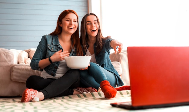 Photo en gros plan d'amies assises sur le sol près du canapé devant un ordinateur portable et elles regardent un film et mangent du pop-corn Des amis drôles se détendent ensemble