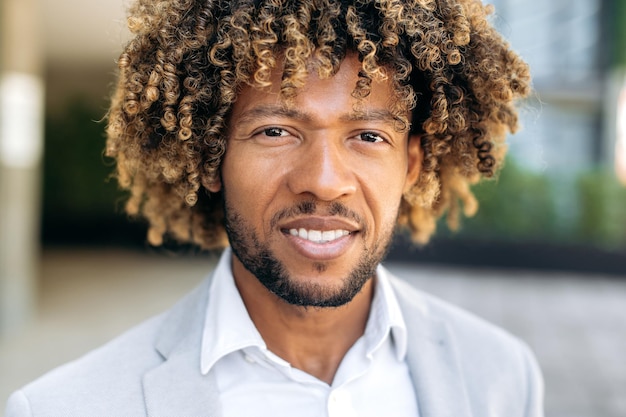 Photo en gros plan d'un beau brésilien charismatique aux cheveux bouclés ou d'un homme hispanique prospère debout à l'extérieur en regardant la caméra en souriant amical